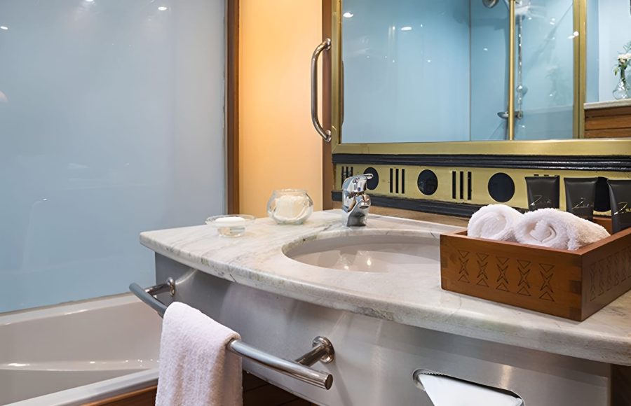A modern bathroom in a cabin on the M/S Alyssa Nile cruise ship, featuring a marble vanity with a recessed sink, a wooden box with rolled white towels, and a bathtub with a frosted glass screen.
