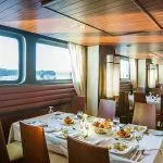 The elegant main restaurant on the M/S Alyssa Nile cruise ship featuring tables set with white linens and gourmet food next to large windows overlooking the Nile River.