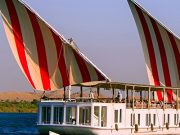 A side view of a white Malouka Nile Dahabiya boat with two large red and white striped sails cruising on the Nile River.