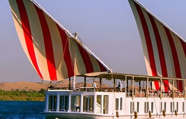 A side view of a white Malouka Nile Dahabiya boat with two large red and white striped sails cruising on the Nile River.