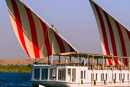 A side view of a white Malouka Nile Dahabiya boat with two large red and white striped sails cruising on the Nile River.
