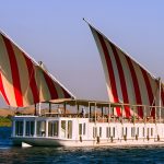 A side view of a white Malouka Nile Dahabiya boat with two large red and white striped sails cruising on the Nile River.
