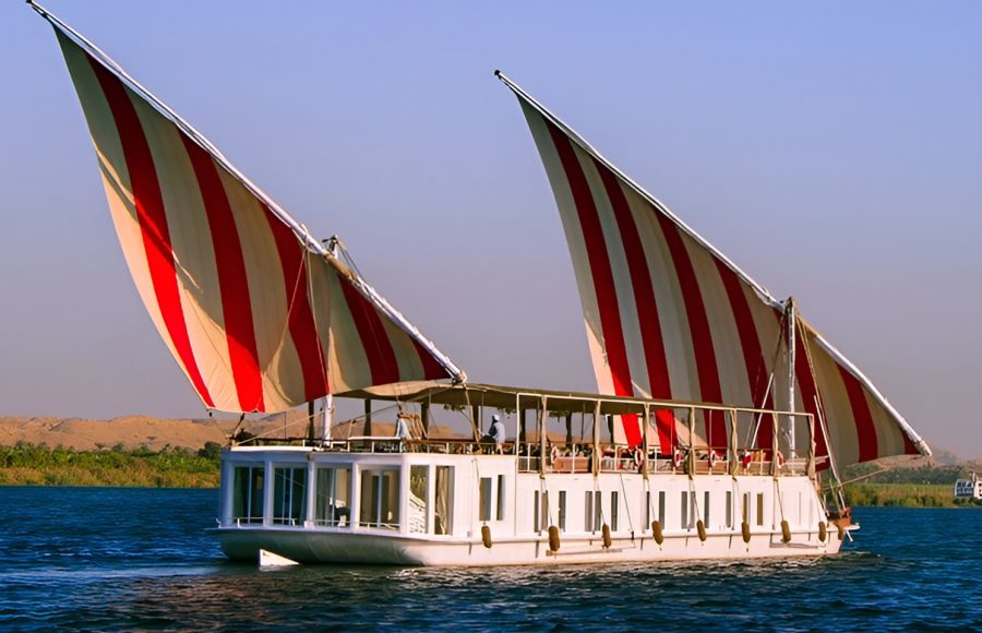 A side view of a white Malouka Nile Dahabiya boat with two large red and white striped sails cruising on the Nile River.