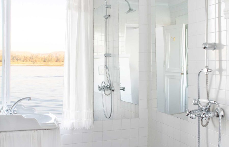 A bright, all-white bathroom on a Nile Dahabiya boat featuring a pedestal sink and shower with a window overlooking the river.