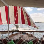 View from the deck of a Dahabiya boat showing red and white striped sails and a striped seating area overlooking the Nile.