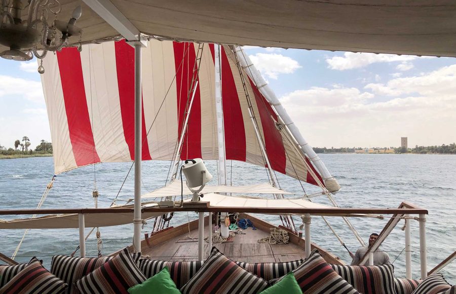 View from the deck of a Dahabiya boat showing red and white striped sails and a striped seating area overlooking the Nile.