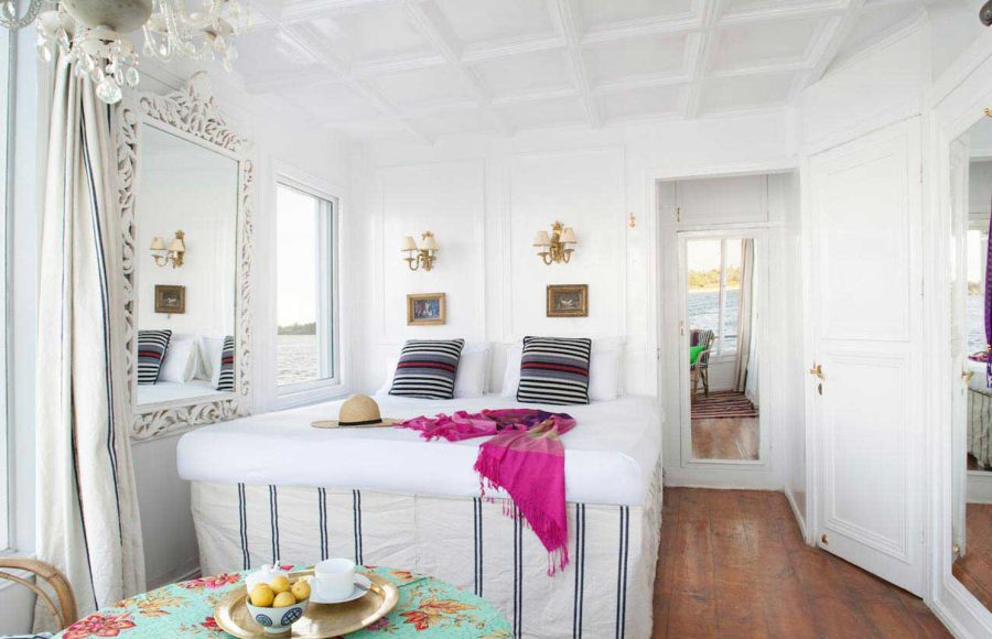 Bright, white panoramic suite on a Nile Dahabiya boat featuring a large bed, striped pillows, and a window view of the river.