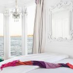 White suite bed with a pink shawl, an ornate mirror, and a crystal chandelier by the window.