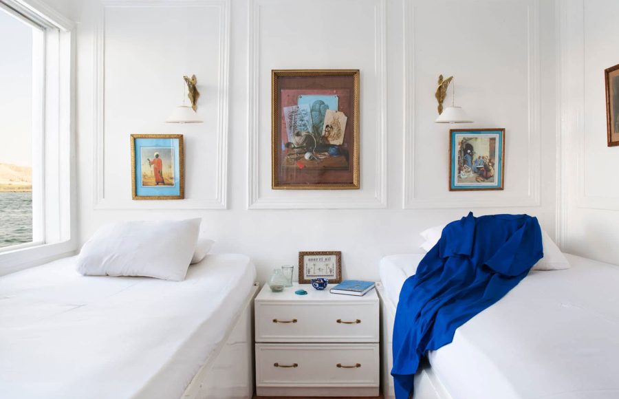 Two white twin beds in a cabin with framed art, wall lamps, and a white nightstand.