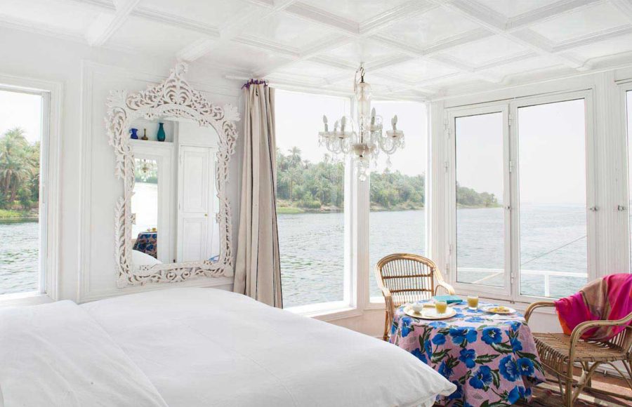 Bright, white luxury cabin on a Nile boat with a bed, ornate mirror, and panoramic windows.