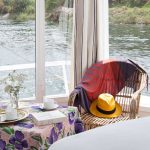 A wicker chair and breakfast table next to a panoramic window on a Nile boat.
