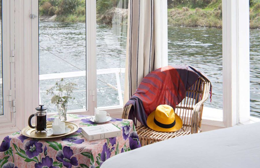 A wicker chair and breakfast table next to a panoramic window on a Nile boat.