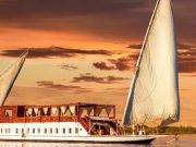 Traditional Egyptian Dahabiya sailing boat on the Nile River at sunset with a sandy riverbank in the background.