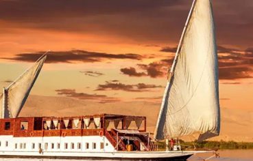 Traditional Egyptian Dahabiya sailing boat on the Nile River at sunset with a sandy riverbank in the background.