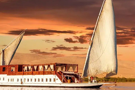 Traditional Egyptian Dahabiya sailing boat on the Nile River at sunset with a sandy riverbank in the background.
