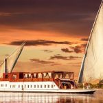 Traditional Egyptian Dahabiya sailing boat on the Nile River at sunset with a sandy riverbank in the background.