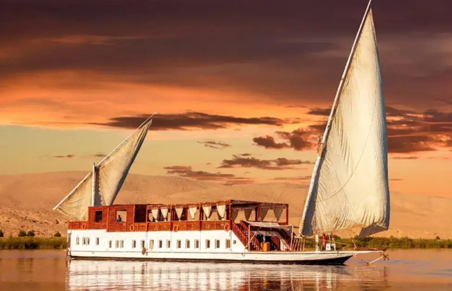 Traditional Egyptian Dahabiya sailing boat on the Nile River at sunset with a sandy riverbank in the background.