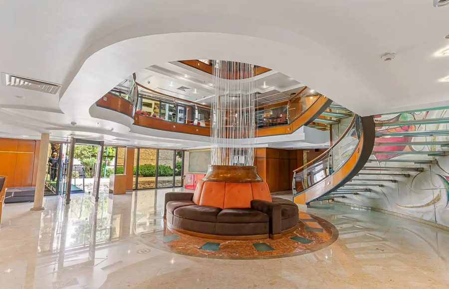 Indoor cruise ship salon with plush seating, central wooden bar, and red accents.