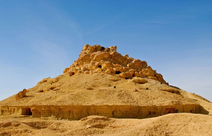 The distinctive, rounded, reddish-brown hill of the Mountain of the Dead in the Siwa Oasis, Egypt, covered in burial holes and tombs, set against a clear blue sky.