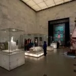 A bright, modern interior gallery of the National Museum of Egyptian Civilization, displaying artifacts in glass cases and featuring a large, ornate textile hanging.