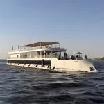 White two-story Dahabiya cruise boat sailing past palm trees.