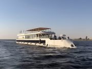 White two-story Dahabiya cruise boat sailing past palm trees.