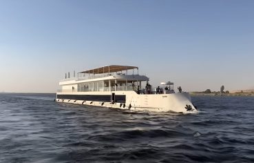 White two-story Dahabiya cruise boat sailing past palm trees.