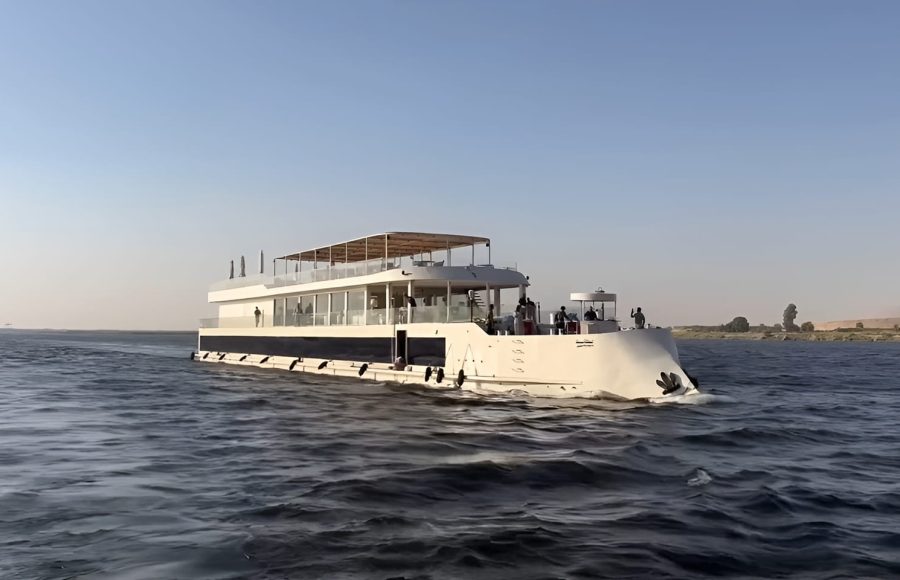 White two-story Dahabiya cruise boat sailing past palm trees.