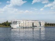 The white, multi-deck exterior of the Nile Crown II cruise ship sailing on the Nile River, featuring a high, pointed bow and rows of windows/balconies, viewed from the water.