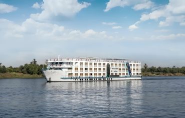 The white, multi-deck exterior of the Nile Crown II cruise ship sailing on the Nile River, featuring a high, pointed bow and rows of windows/balconies, viewed from the water.