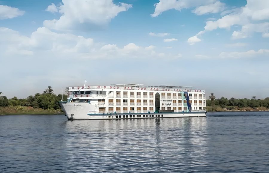 The white, multi-deck exterior of the Nile Crown II cruise ship sailing on the Nile River, featuring a high, pointed bow and rows of windows/balconies, viewed from the water.