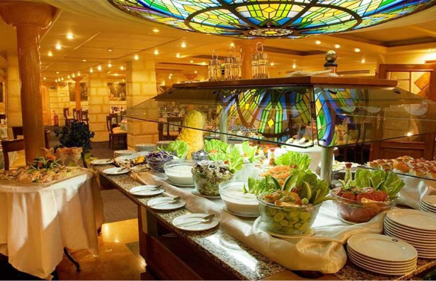 A lavish buffet station inside the Nile Crown II restaurant, displaying fresh salads, appetizers, and stacks of plates, beneath a brightly lit, circular stained-glass ceiling dome.