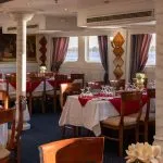 Interior of the Nile Crown II restaurant with set dining tables, featuring white tablecloths, red runners, wooden chairs, arched windows overlooking the Nile River, and a large classical painting.
