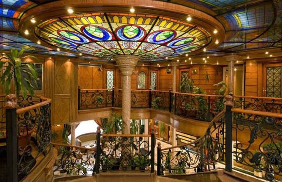 A lavish multi-story lobby on the Nile Crown II cruise ship, featuring a central pillar, elaborate wooden banisters, wrought-iron railings, potted plants, and a stunning circular stained-glass ceiling dome.