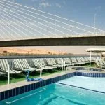Two set dinner tables with bottles of wine and chairs on the sundeck of the Nile Crown II cruise ship, overlooking an illuminated, tiered hill and the Nile River at twilight.