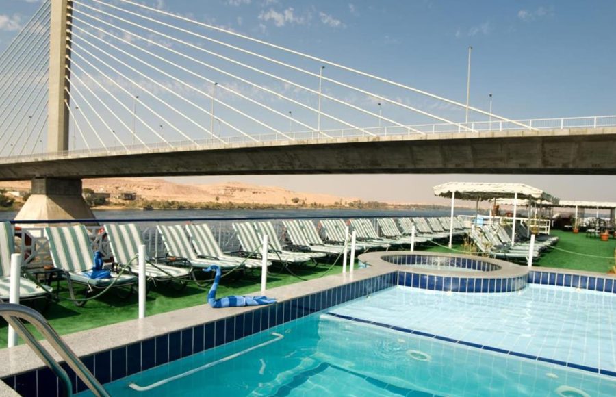 Two set dinner tables with bottles of wine and chairs on the sundeck of the Nile Crown II cruise ship, overlooking an illuminated, tiered hill and the Nile River at twilight.