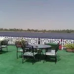 Outdoor seating area on the sundeck of the Nile Crown II cruise ship with tables, chairs, and potted plants, overlooking the Nile River and a distant tree-lined bank under a hazy sky.