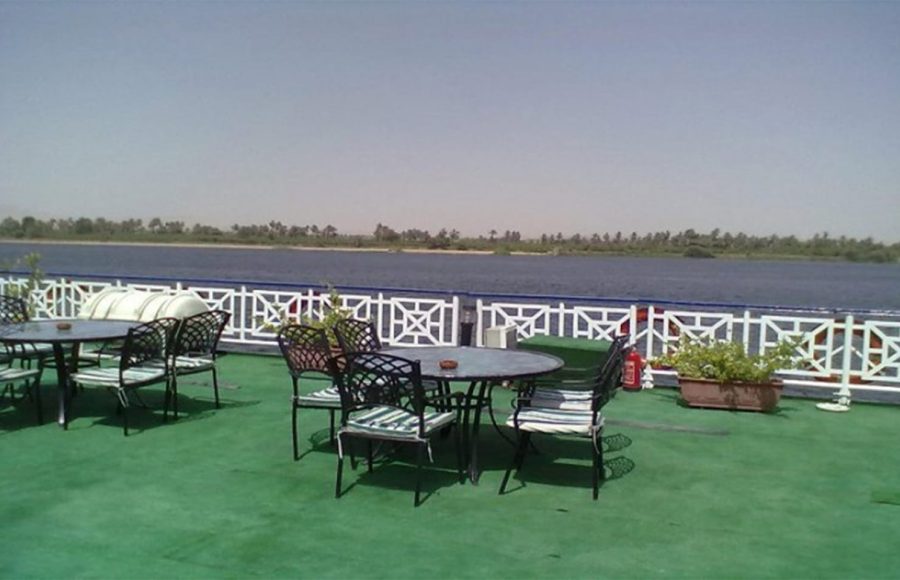 Outdoor seating area on the sundeck of the Nile Crown II cruise ship with tables, chairs, and potted plants, overlooking the Nile River and a distant tree-lined bank under a hazy sky.