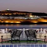 Two set dinner tables and chairs on the sundeck of the Nile Crown II cruise ship, overlooking a dramatically lit, tiered hill and the Nile River at twilight.