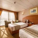 A bright twin-bedded cabin on the Nile Crown II cruise ship, featuring two beds with striped linens, a wood headboard, and a large sliding glass door leading to a balcony.