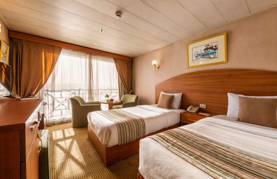 A bright twin-bedded cabin on the Nile Crown II cruise ship, featuring two beds with striped linens, a wood headboard, and a large sliding glass door leading to a balcony.
