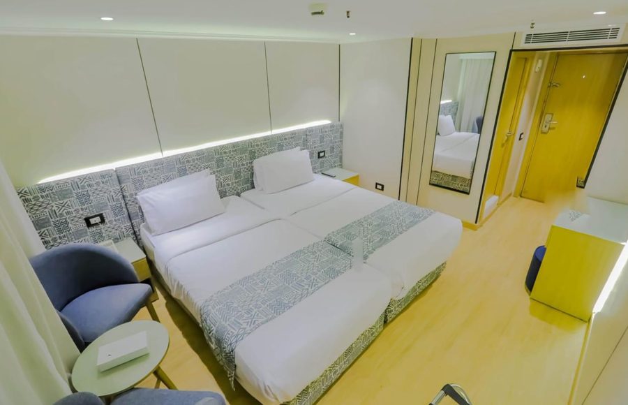 A bright, modern twin cabin on the Nile Palace cruise ship with light wood floors, two single beds, and a full-length mirror, featuring a blue and white patterned headboard and bed runners.