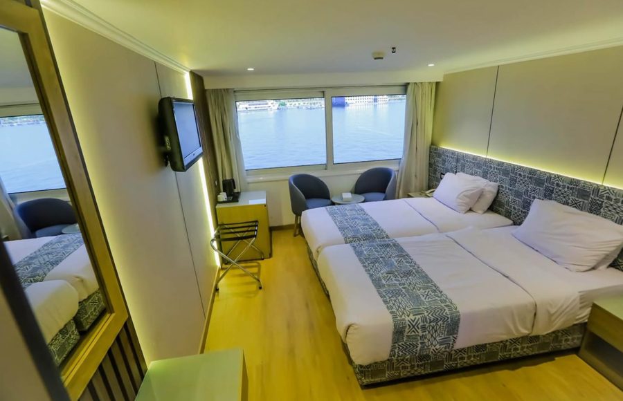 A contemporary twin cruise cabin with light wood floors, two single beds with patterned runners, a full-length mirror, and large windows with a Nile River view.