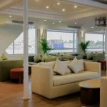 A spacious cruise ship lounge or bar area with light wood floors, white structural columns, and numerous seating areas with beige and light olive green sofas and windows overlooking the Nile.