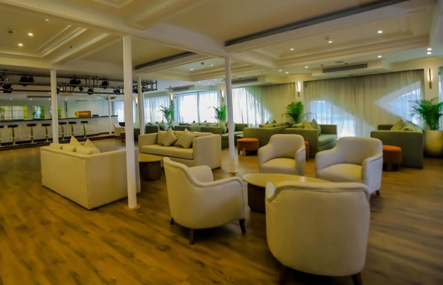 A spacious cruise ship lounge with light wood floors, an inner bar area visible in the background, and various seating groups with cream armchairs and olive-green sofas.