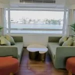 A close-up view of a seating group in the Nile Palace Deluxe lounge, featuring two sage green sofas, modern gold and white coffee tables, and large windows with a clear view of the city shoreline across the Nile River.