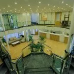 A two-story cruise ship lobby and reception area viewed from the top of the stairs, featuring white walls, light wood flooring, glass railings, and a central reception desk.