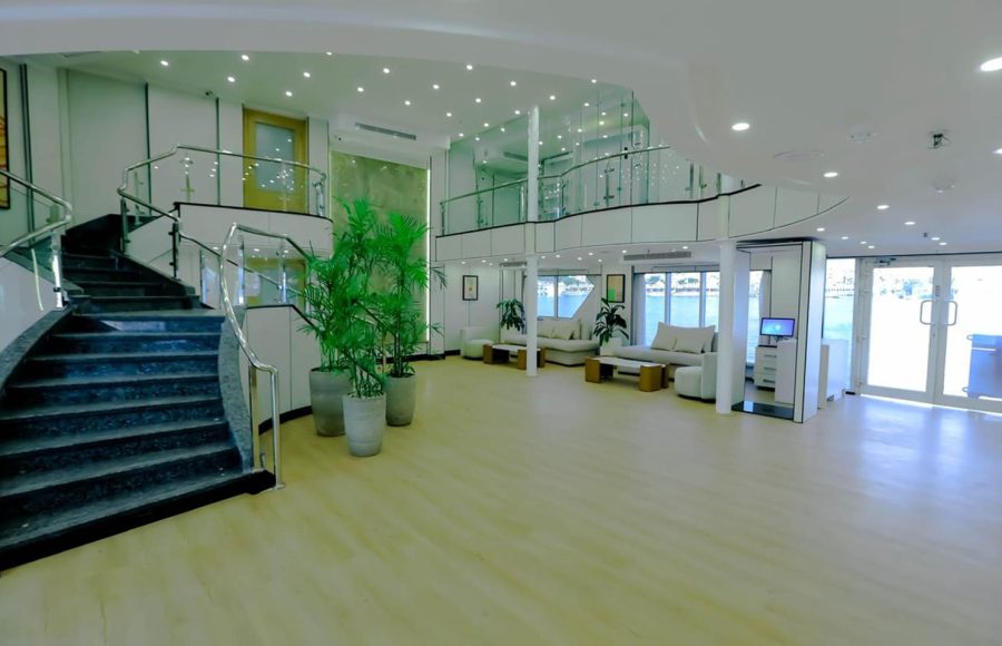 A brightly lit, modern lobby on the Nile Palace Deluxe cruise ship. It features a curved staircase with dark steps and a glass railing, light wood flooring, several comfortable white seating areas, and large potted plants. In the background, there is an upper mezzanine level also with a glass railing, and glass doors leading outside to the water.