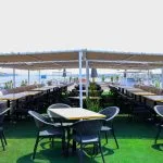The Nile Palace Deluxe sun deck dining area, featuring rows of black wicker chairs and small tables on green artificial turf, covered by a large beige awning, with a view of the river and city skyline.