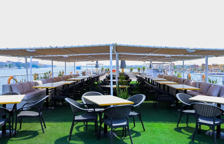 The Nile Palace Deluxe sun deck dining area, featuring rows of black wicker chairs and small tables on green artificial turf, covered by a large beige awning, with a view of the river and city skyline.